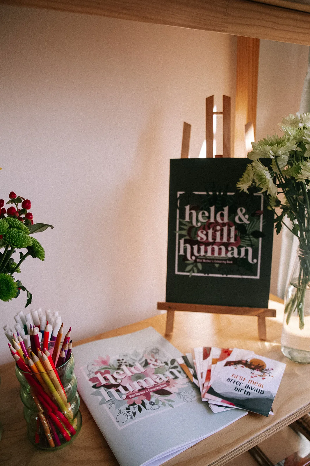 The Held & Still Human colouring book on an easel, with colouring pencils and a stack of milestone cards