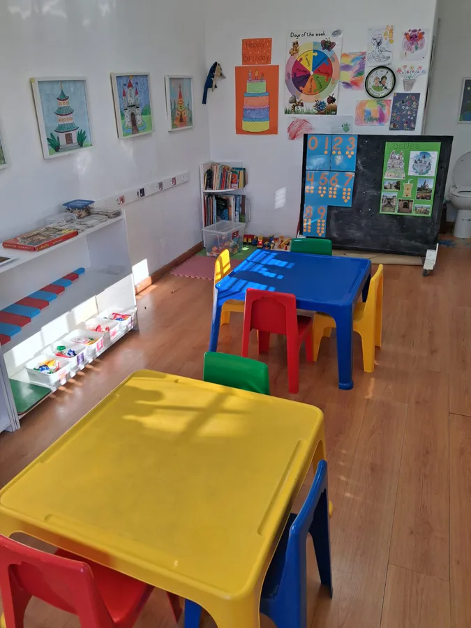 The classroom, with child-height tables and chairs, Montessori materials on low open shelves and children's artwork on the walls
