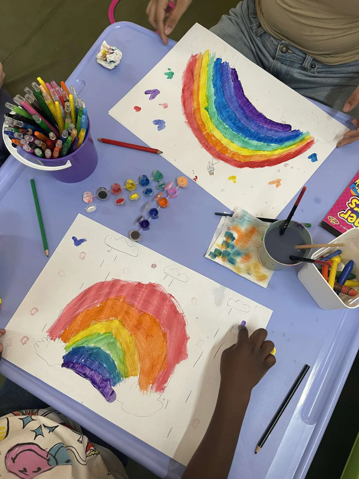 Two children's rainbow paintings on a purple table with paints and pencils
