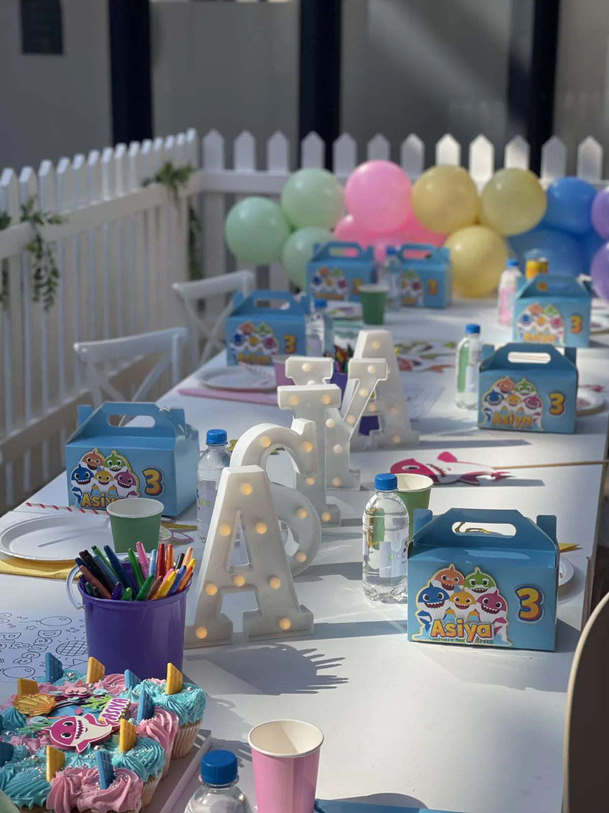 A party table set with balloons, themed party boxes and cupcakes behind a white picket fence