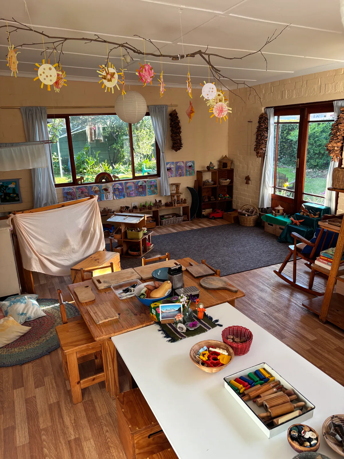 The Rainbow Nursery School playroom — wooden tables and stools, low shelves of natural toys, a play tent and paper suns hung from a branch across the ceiling